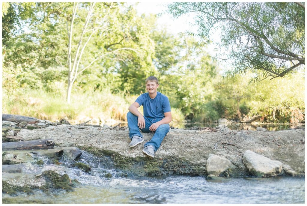 senior boy sitting on a large rock next to a creek outside