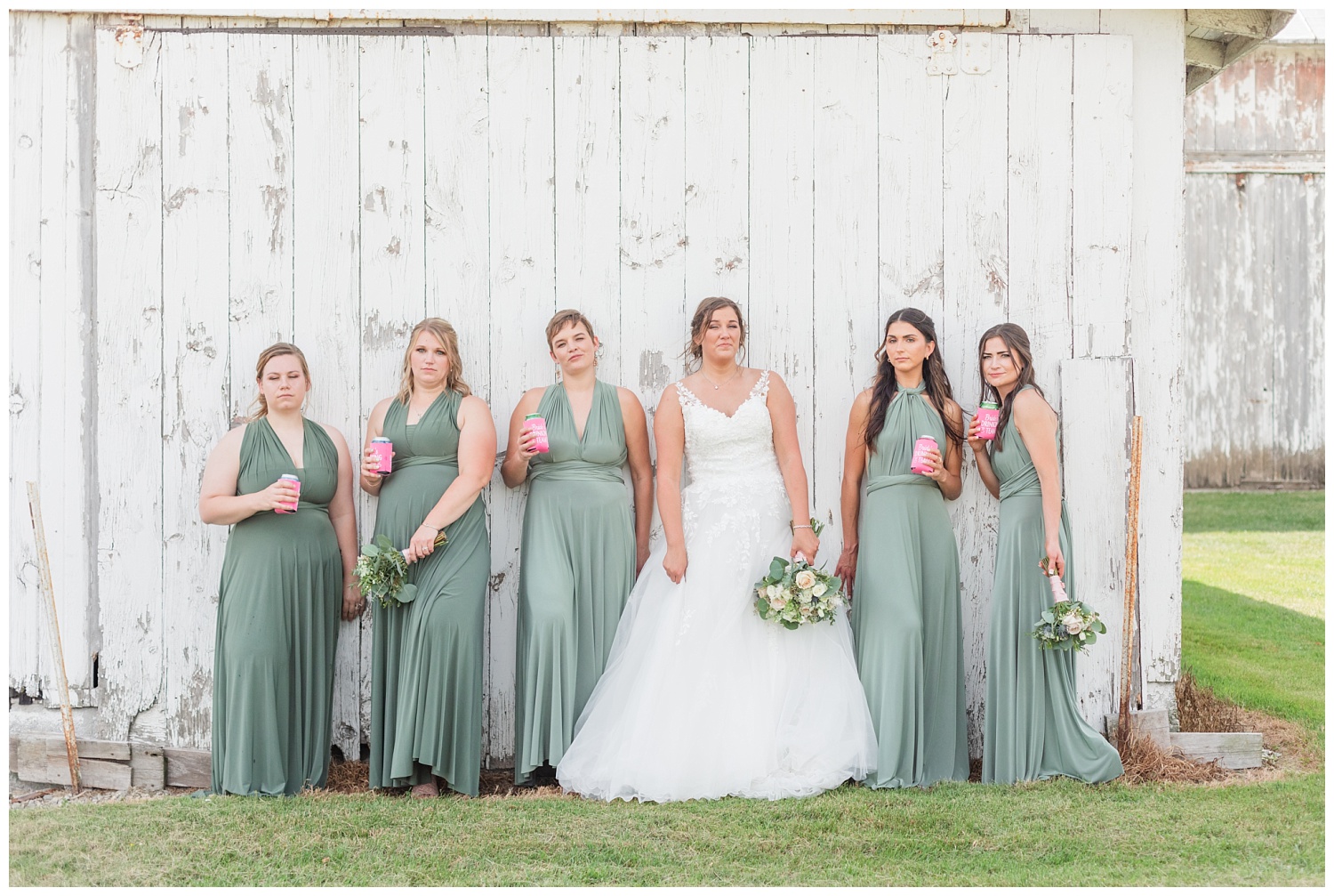 bridal party posing with beers against a white barn