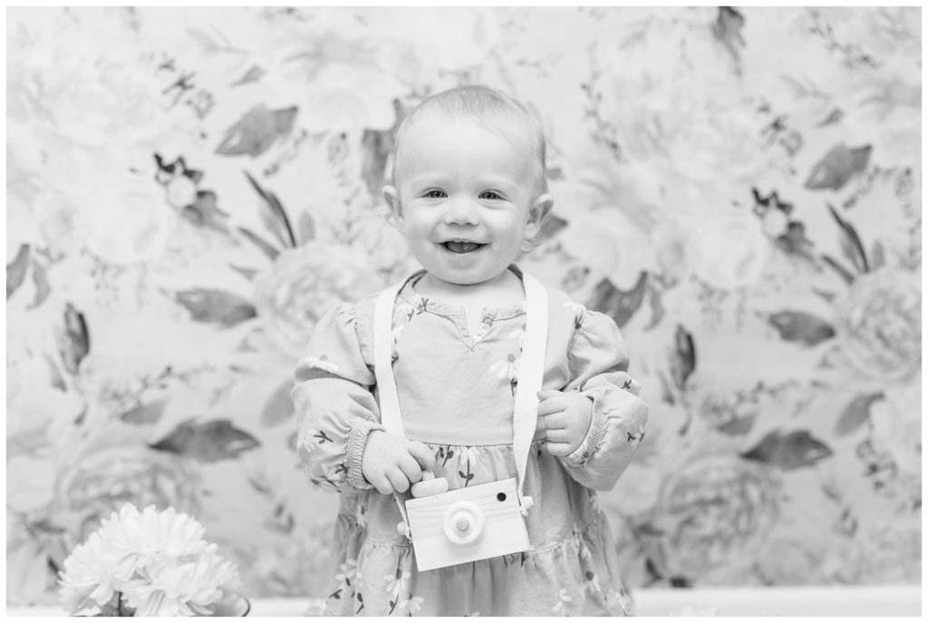 little girl smiling and wearing a wooden camera at birthday photo session