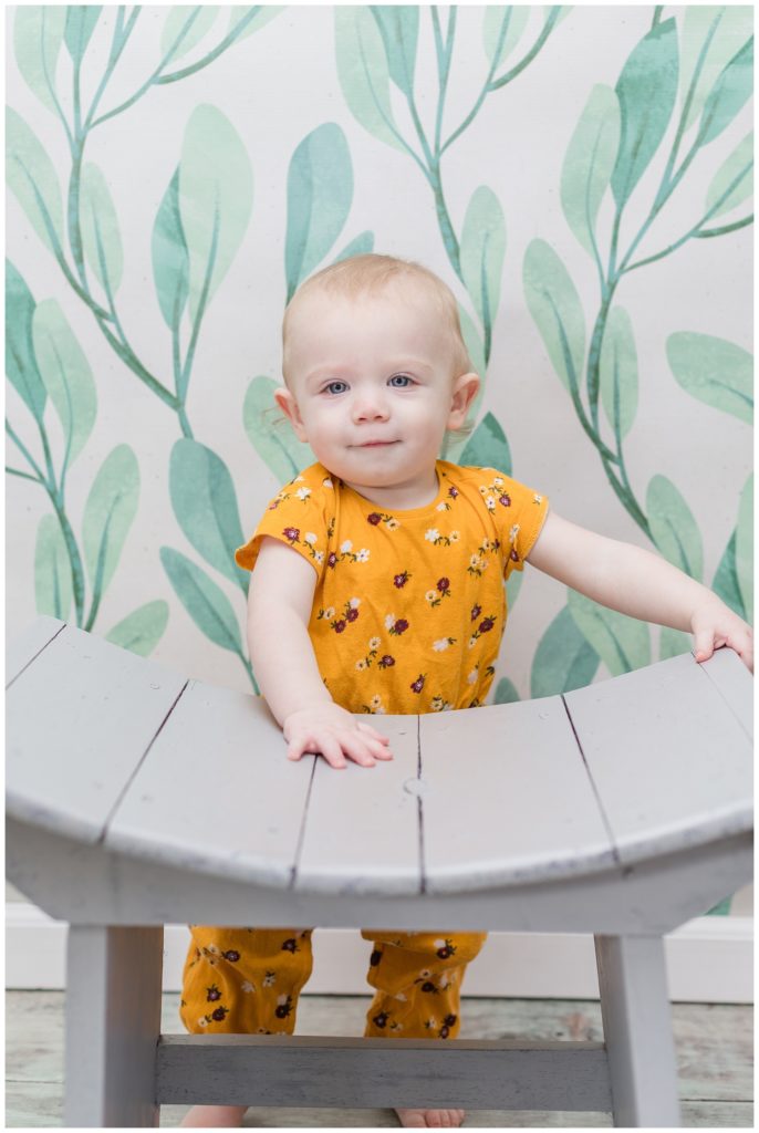 one year old girl wearing a yellow romper standing behind a gray bench