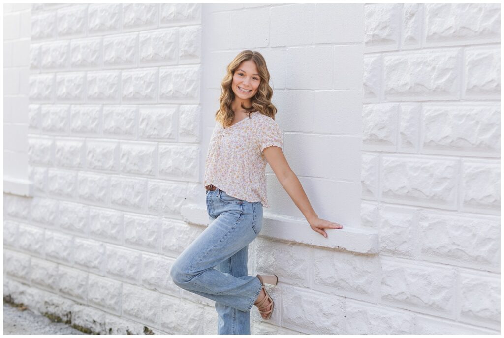 senior wearing a top and jeans leaning against a white wall in Ohio