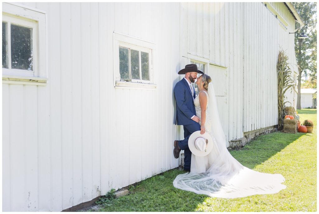 bride and groom leaning against white barns amid fall decorations