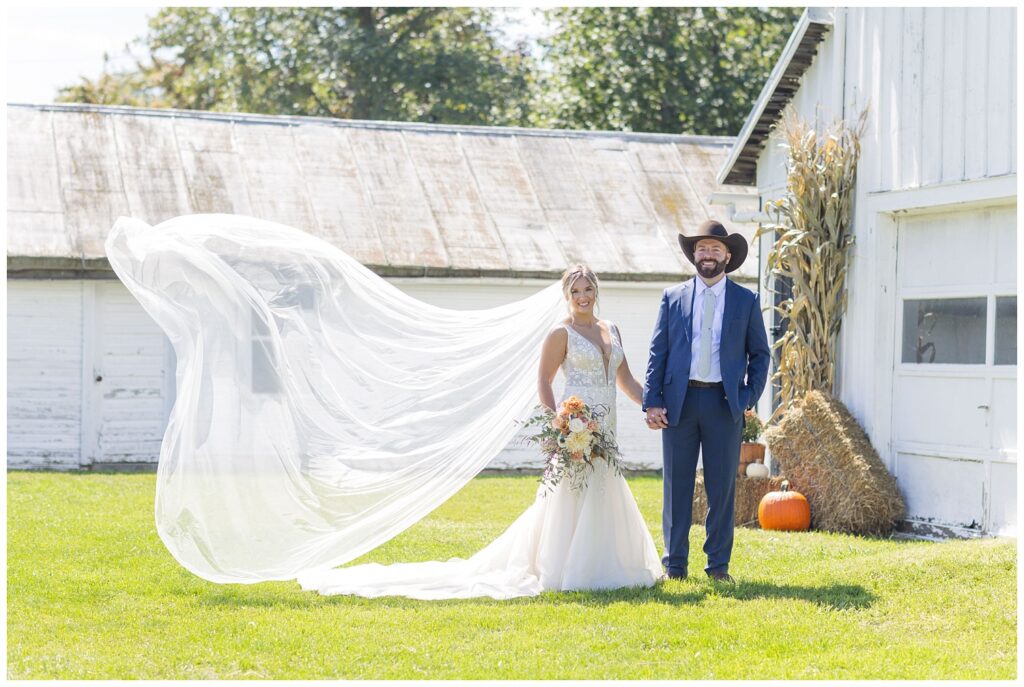 bride's veil flying in the wind while couple holds hands next to a white barn