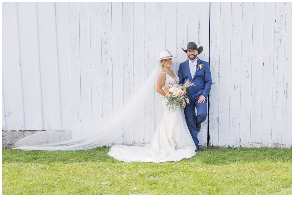 bride and groom wearing cowboy hats leading against a white barn