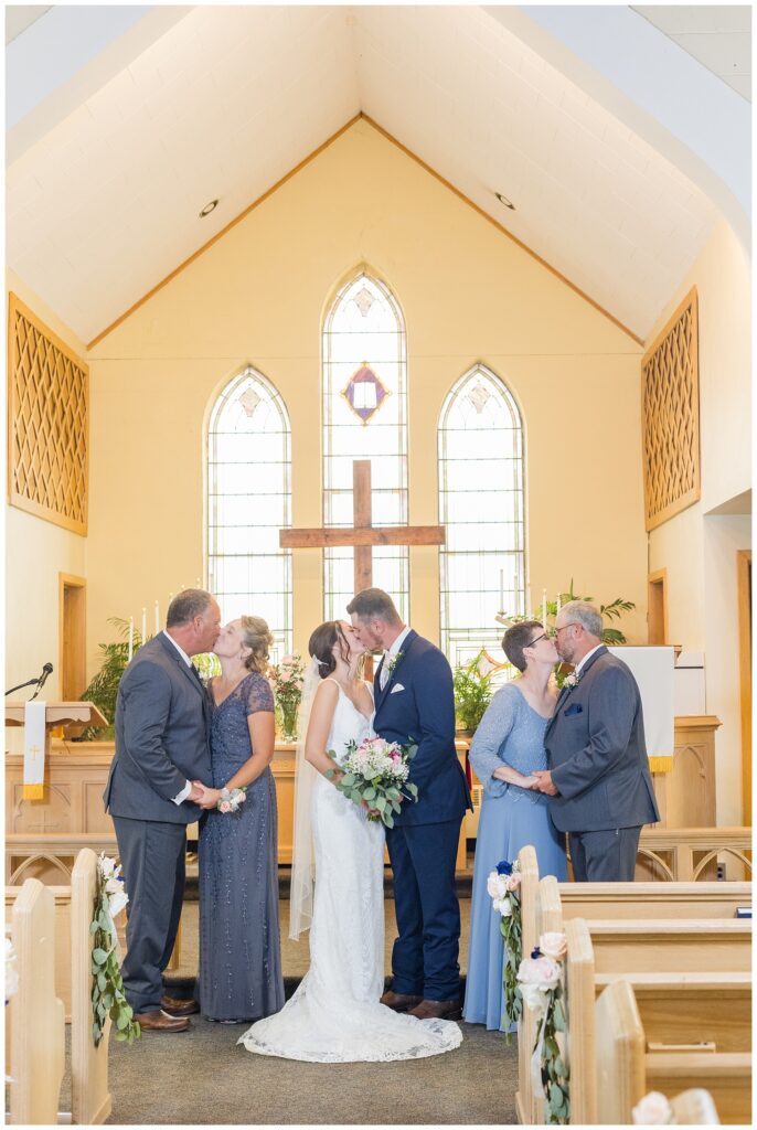 bride and groom, bride's parents and groom's parents all share a kiss at church