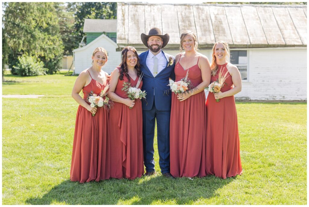 groom posing with bridesmaids in front of a white barn