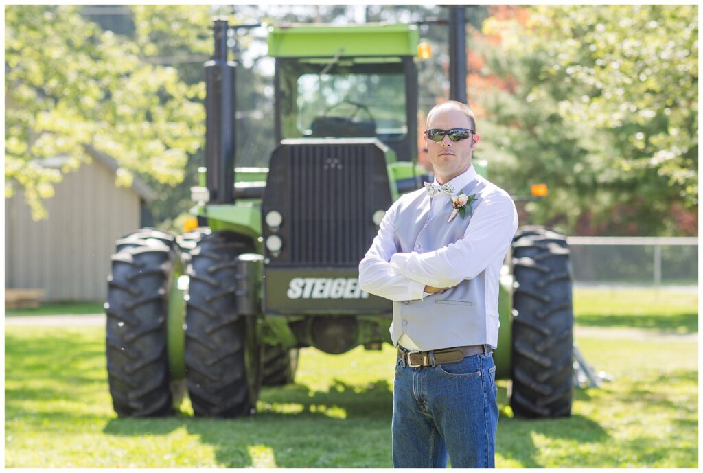 groom wearing sunglasses and posing in front of a large green tractor 