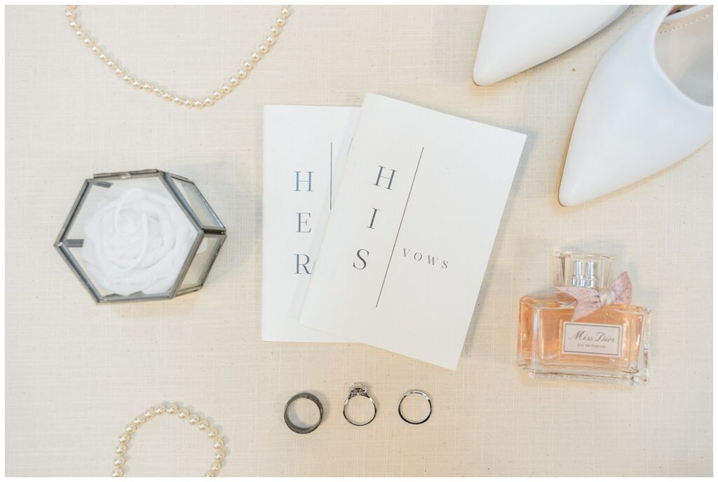 white wedding shoes, vow books and perfume sitting on a ivory mat 