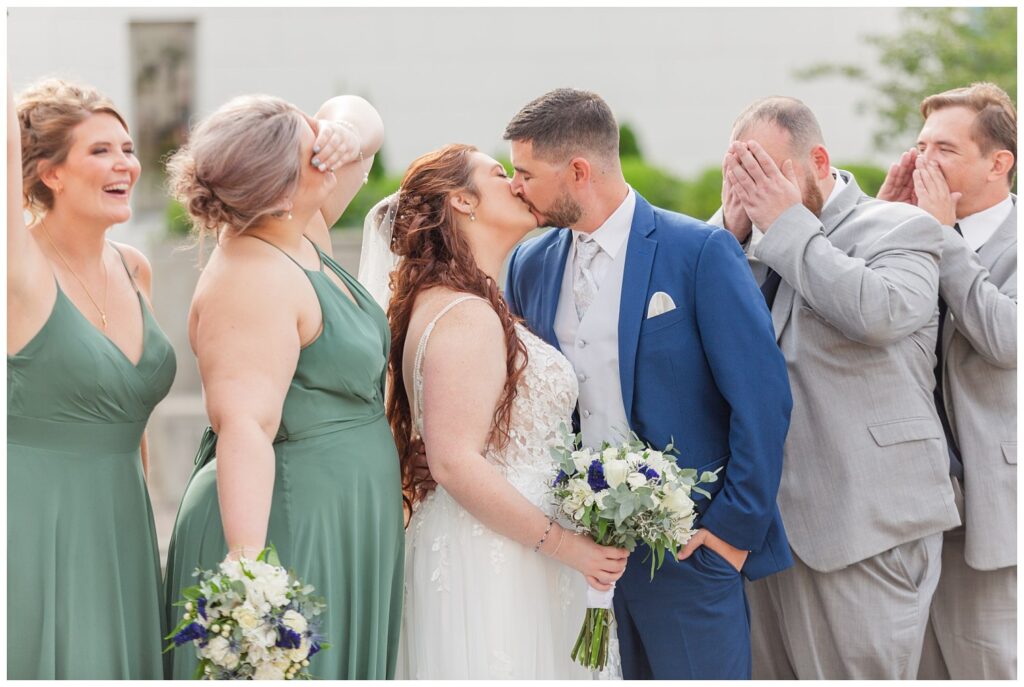 bride and groom share a kiss while the wedding party clooes their eyes