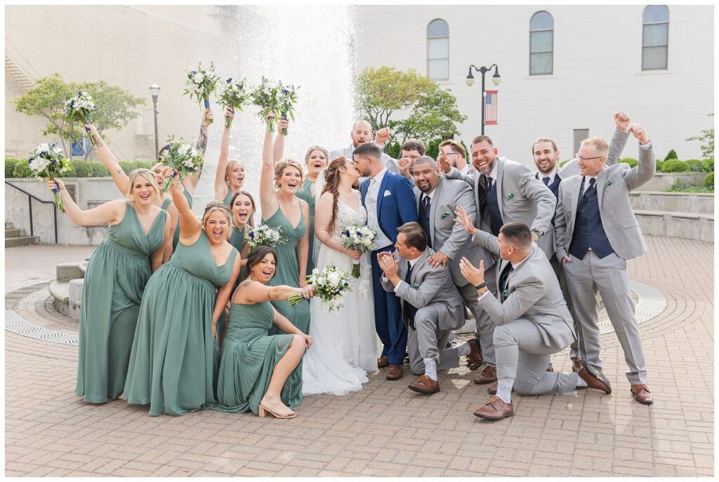 full wedding party cheering for bride and groom as they kiss in front of the fountain