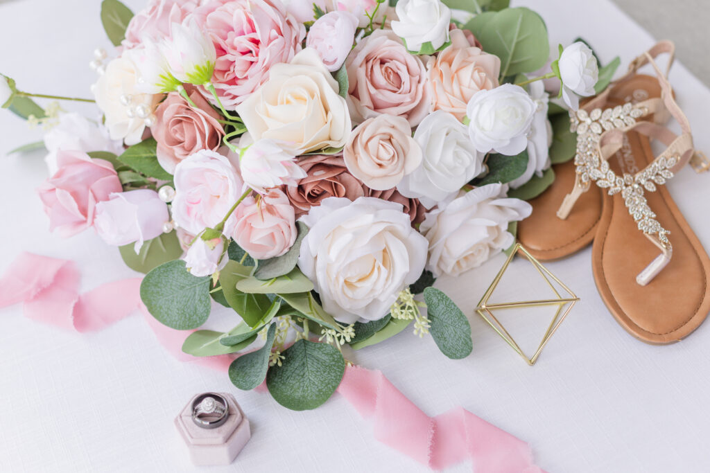 floral bouquet sitting on top of a white mat and pink ribbon next to sandals and ring box