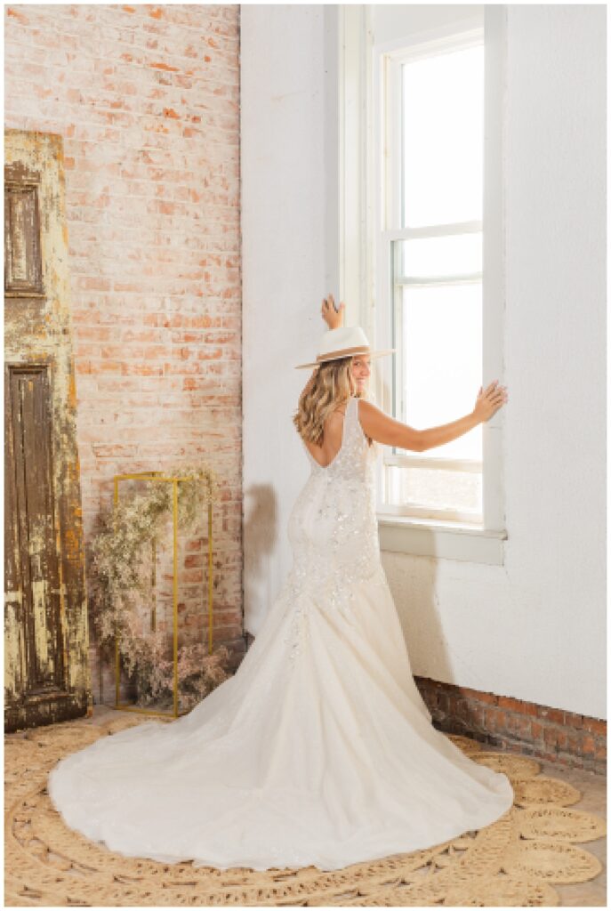 bride posing in front of a window in a brick room wearing her dress and cowboy hat