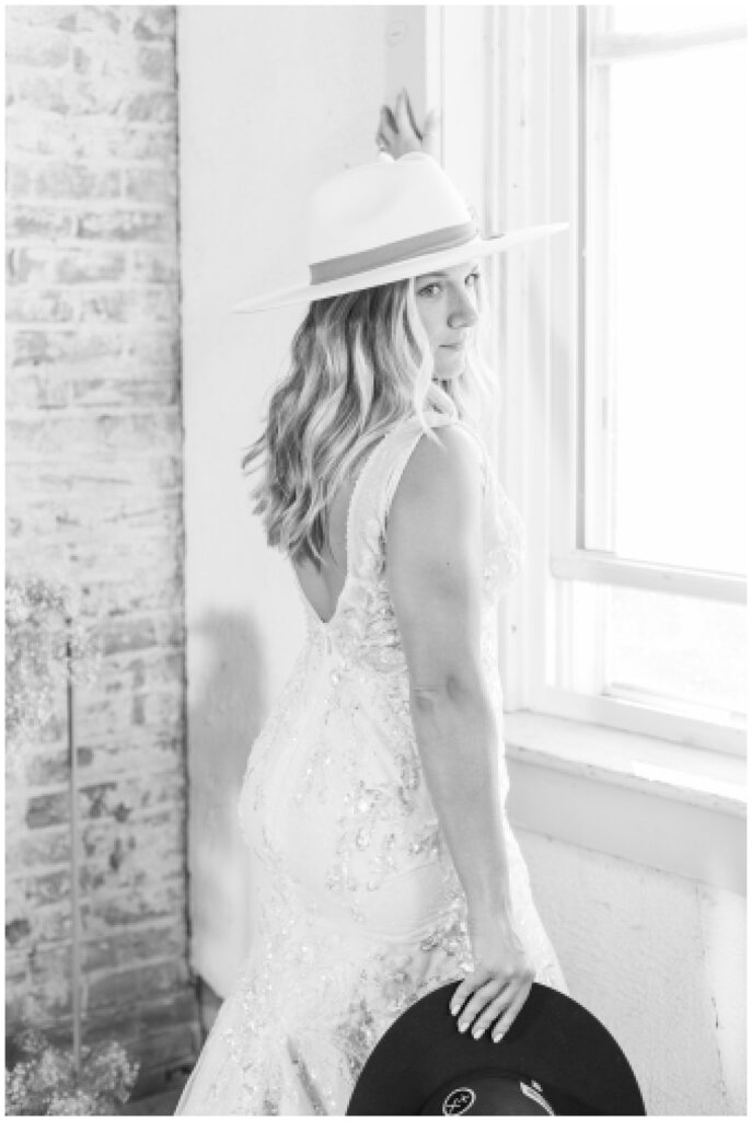 bride standing in front of a window wearing an ivory cowboy hat and holding the groom's hat