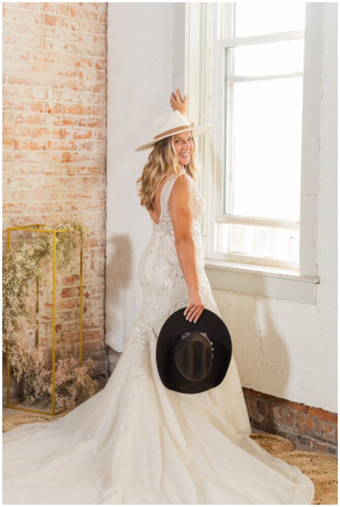 bride posing in front of a window holding the groom's black cowboy hat