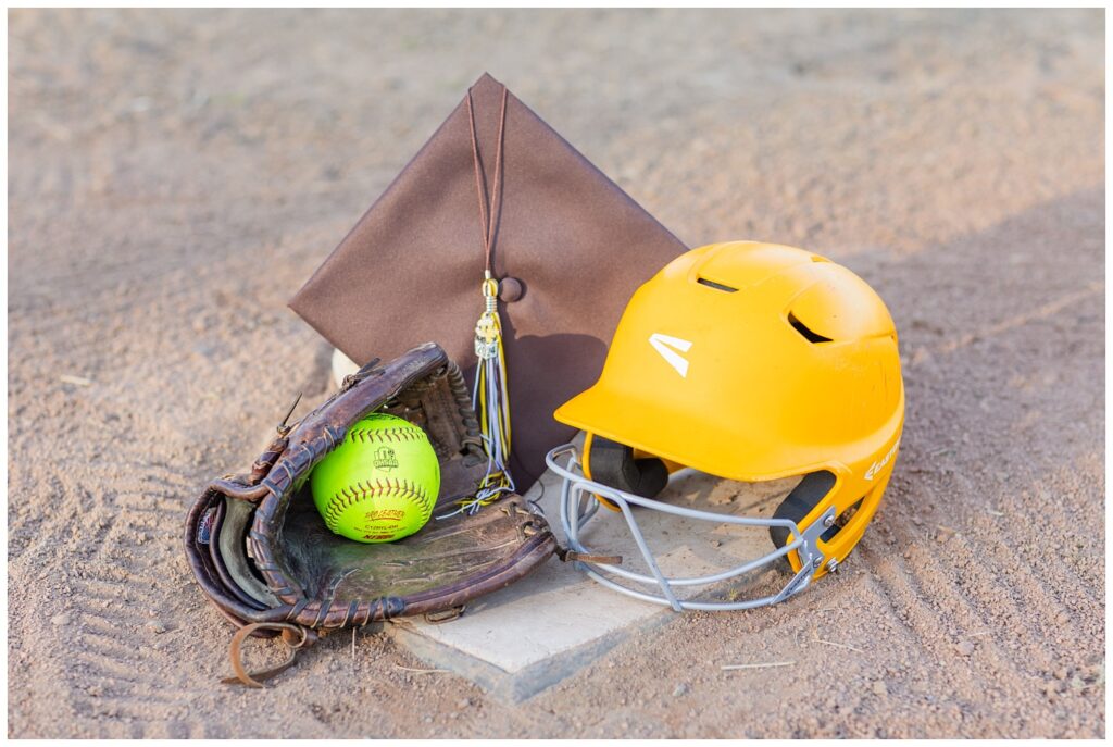 brown cap, softball and glove, and yellow helmet sitting on a base