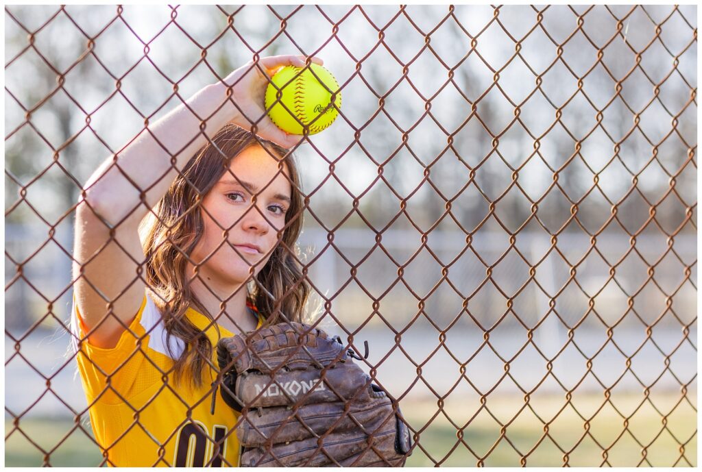 senior girl leaning against a fence holding a glove and softball