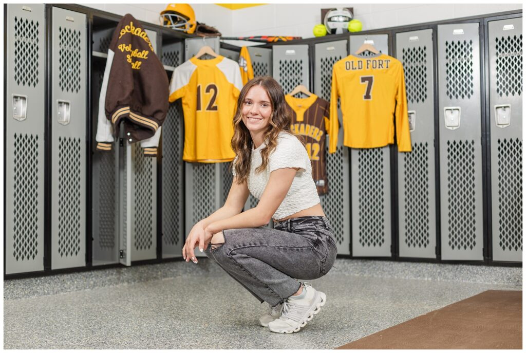 senior girl squatting in a locker room with all of her sports uniforms in Old Fort High School