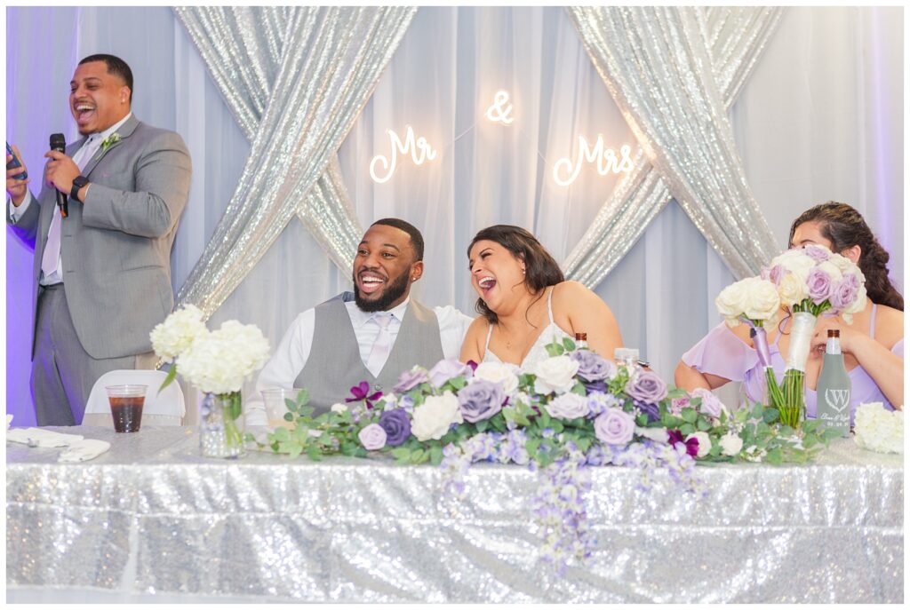 bride and groom laughing at wedding reception while groomsmen is reading toast
