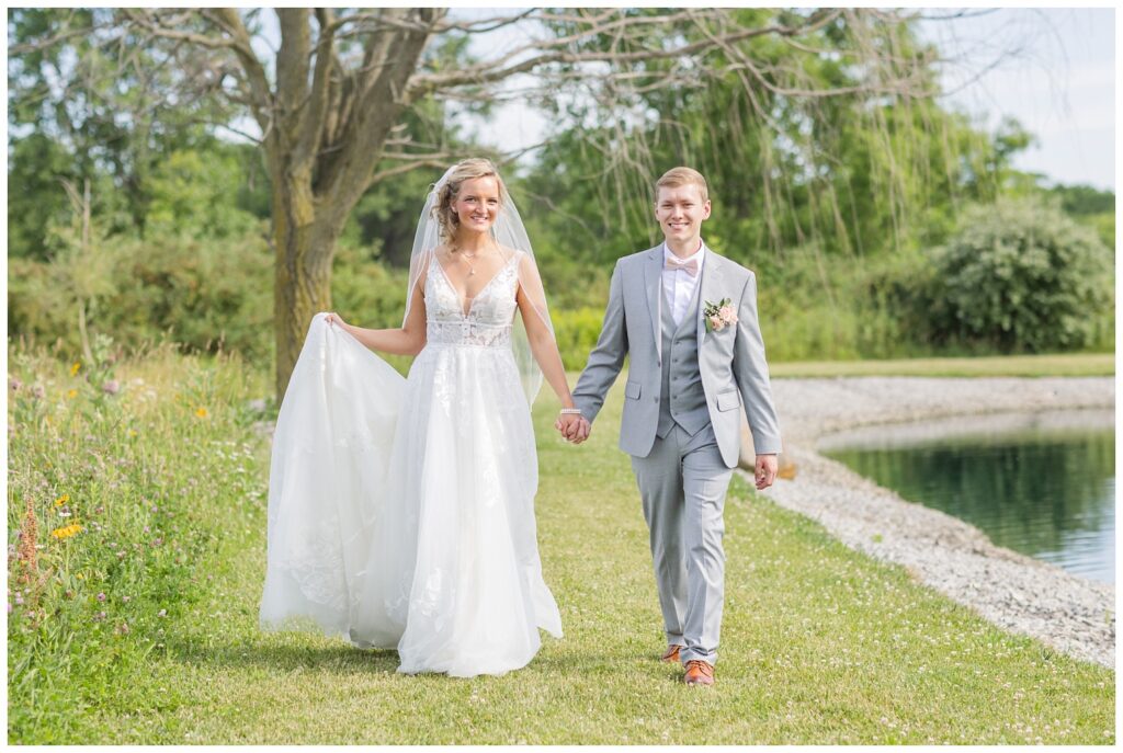 bride and groom walking along the side of the pond while bride is holding her dress