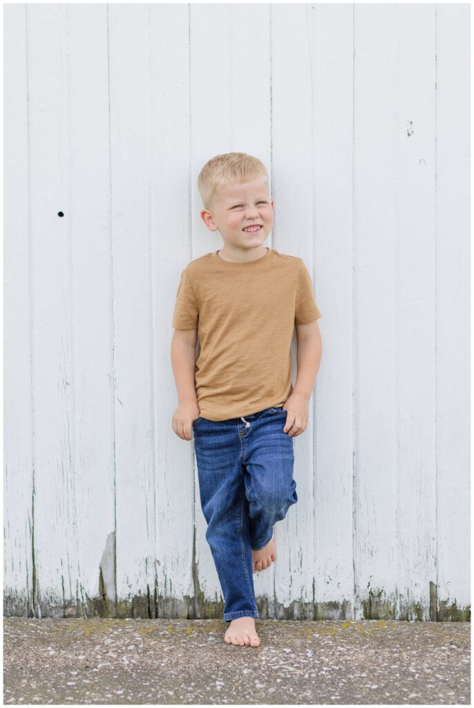 little boy wearing a tan shirt and jeans posing against a white barn in Tiffin, Ohio