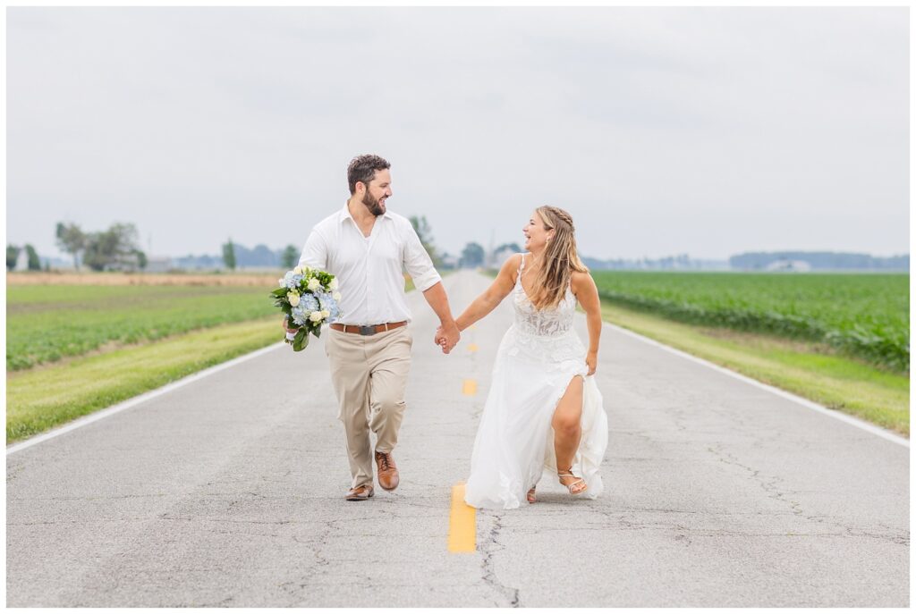 bride and groom running and holding hands in the middle of a road next to a field 