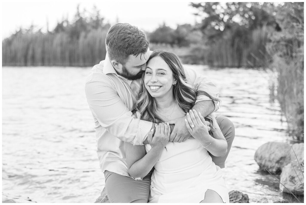 man hugging his fiance from behind while sitting on a rock next to a lake