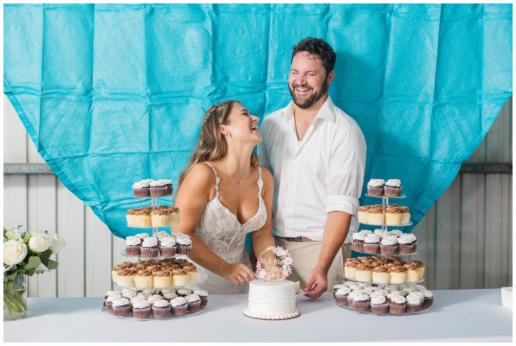 groom and bride laughing while cutting their cake in front of a blue decorations in family barn