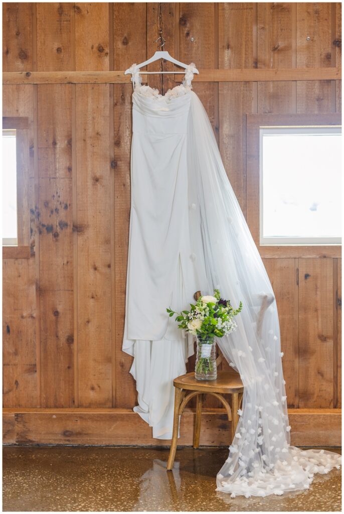 white wedding dress hanging from a wooden wall with veil draped over a chair