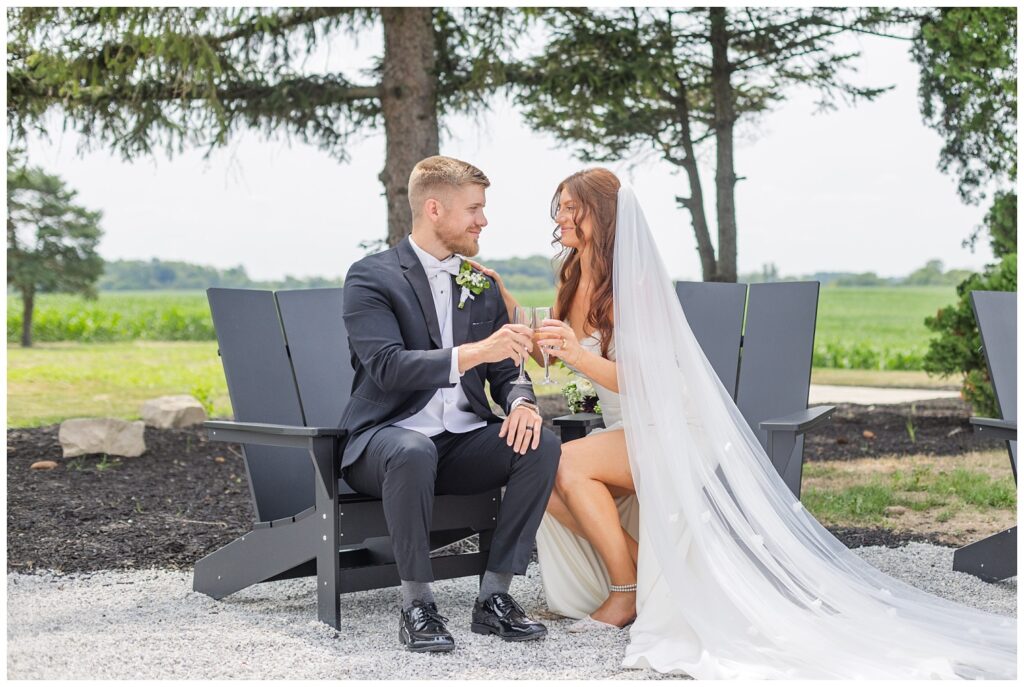 bride and groom holding champagne glasses and sitting in black Adirondack chairs on the venue's patio