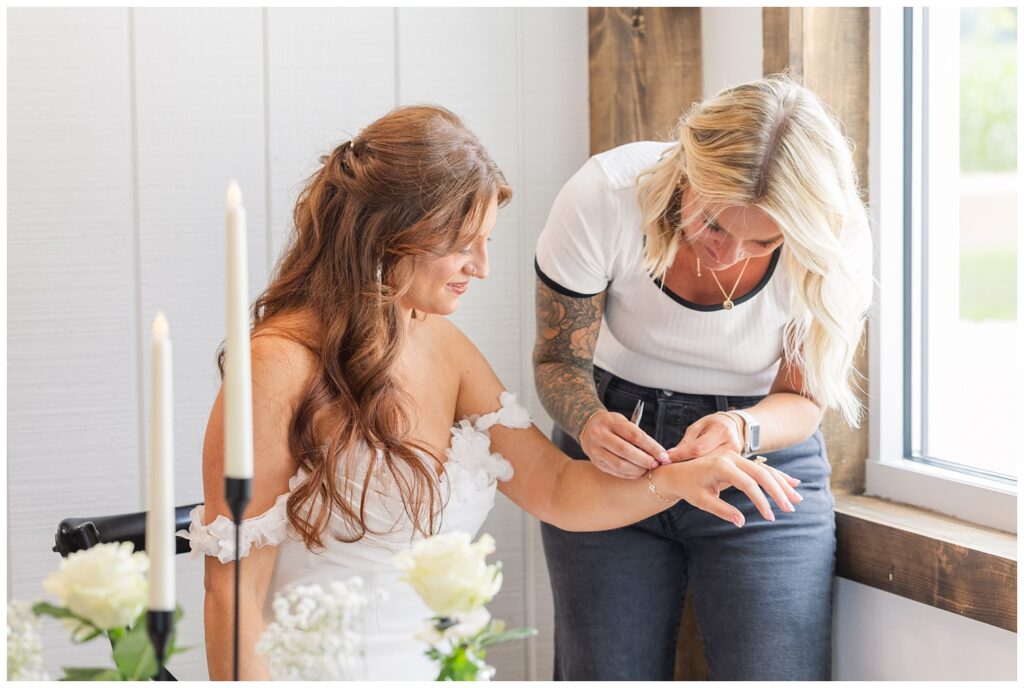 bride getting permanent jewelry put on next a window at wedding venue