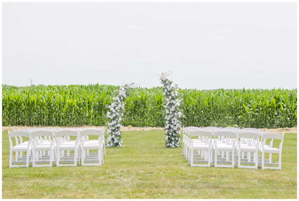 ceremony site with white chairs and floral arch set up in front of a corn field