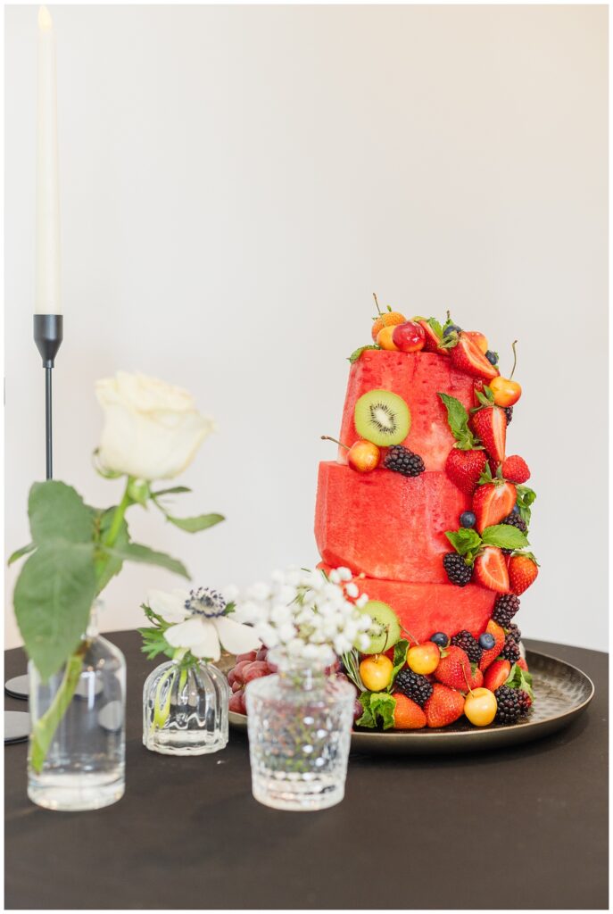 tall watermelon fruit shaped cake on a table next to glass vases filled with white flowers