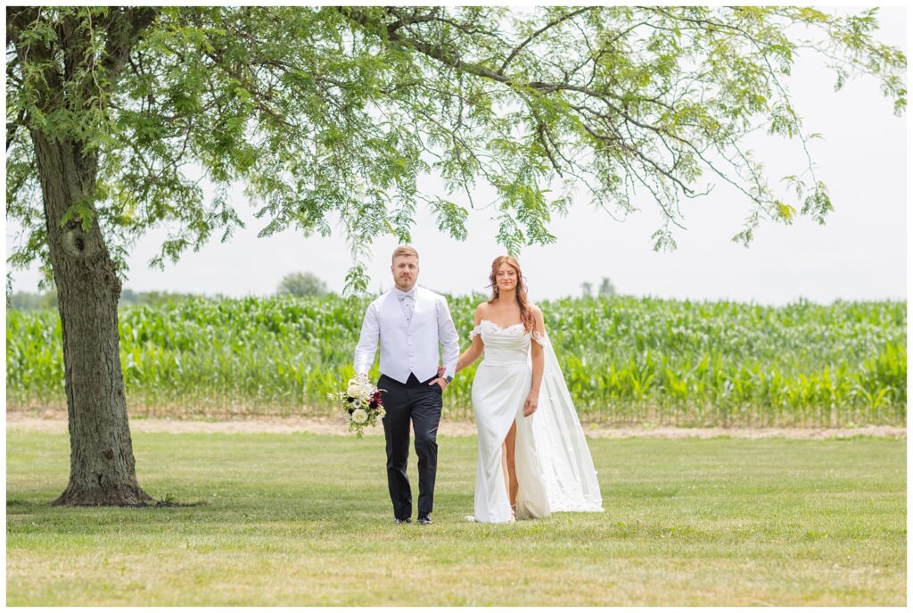 groom holding the bride's bouquet while posing in front of a corn field 