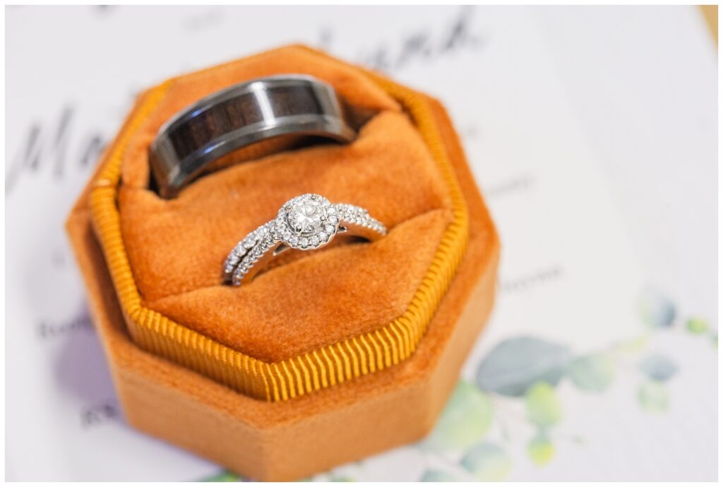 wedding rings sitting in an orange ring box on top of an invitation 