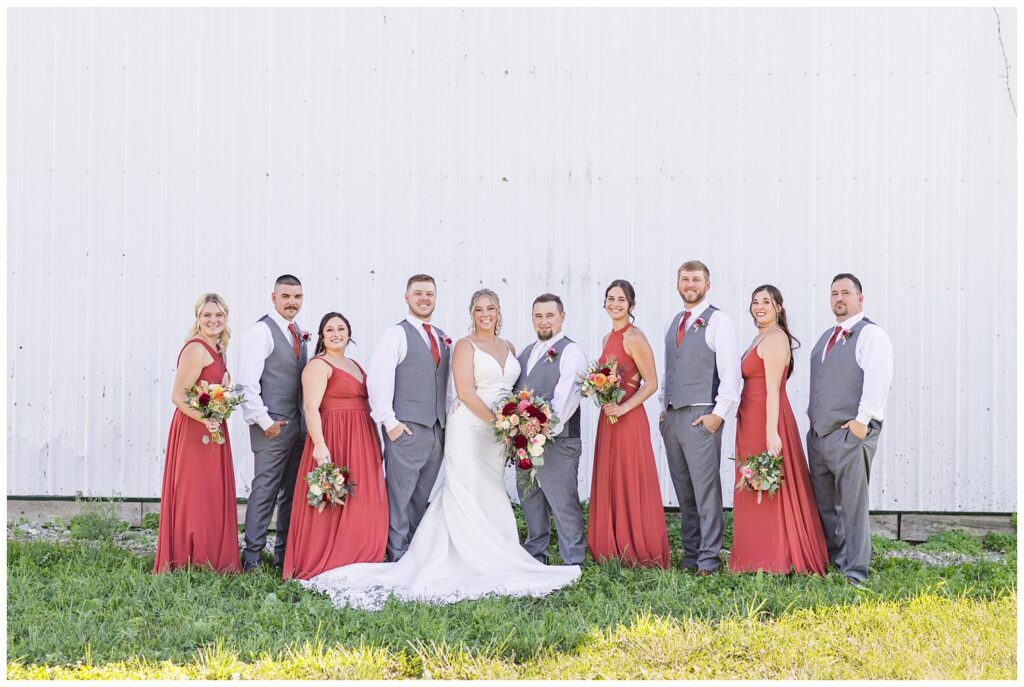 full wedding party posing in front of a white wall of a barn on a dairy farm