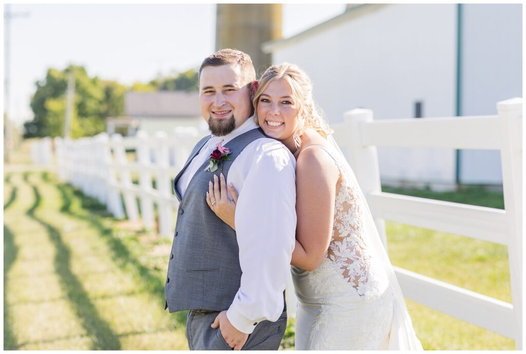 bride hugging the groom groom behind next to a large white fence