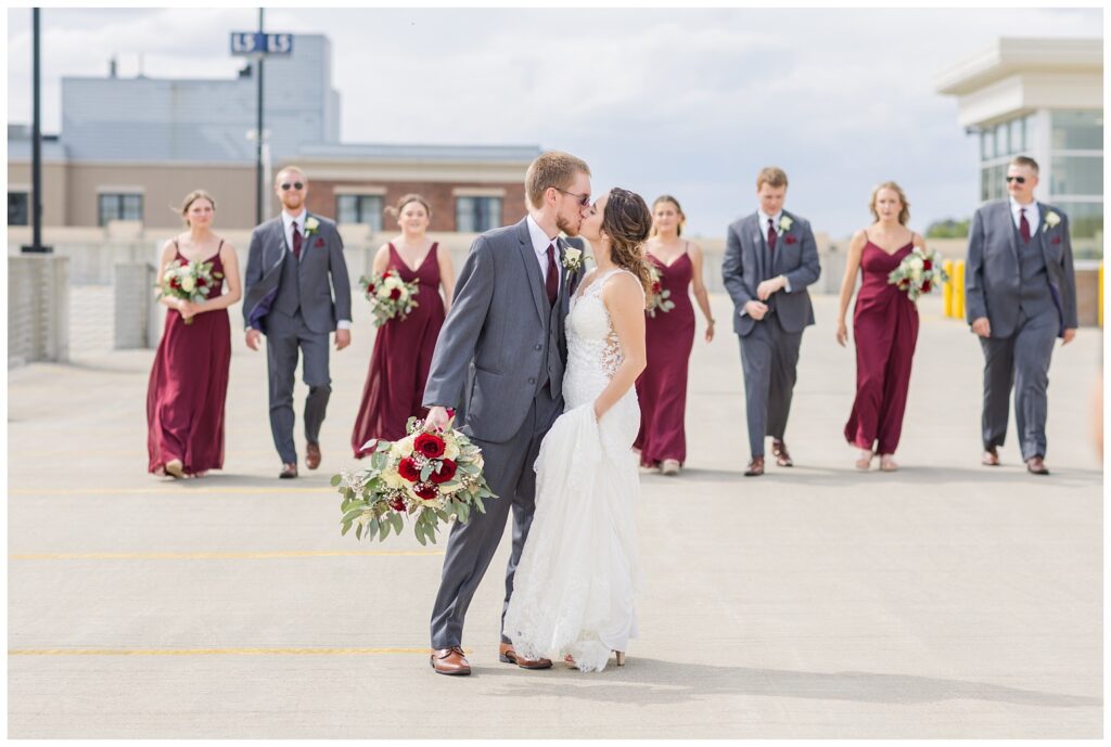 bride and groom share a kiss while the wedding party is walking behind them