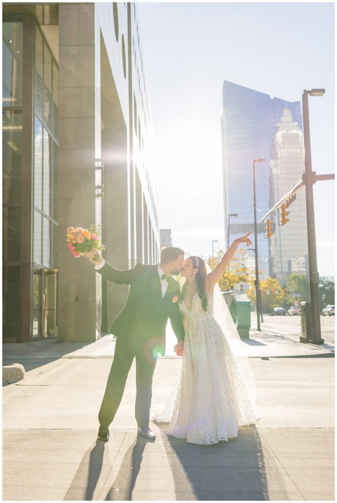 groom holding the bride's bouquet while they kiss on the sidewalk outside