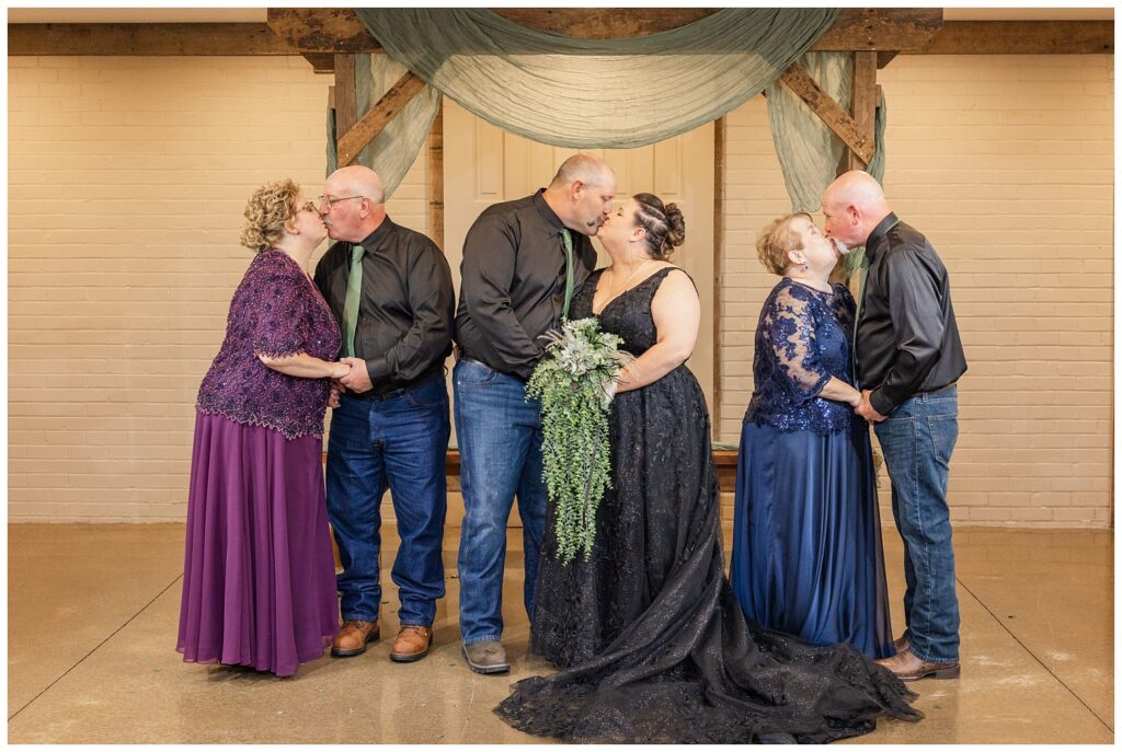 bride and groom and both sets of their parents all share a kiss under the wooden overhand inside chapel