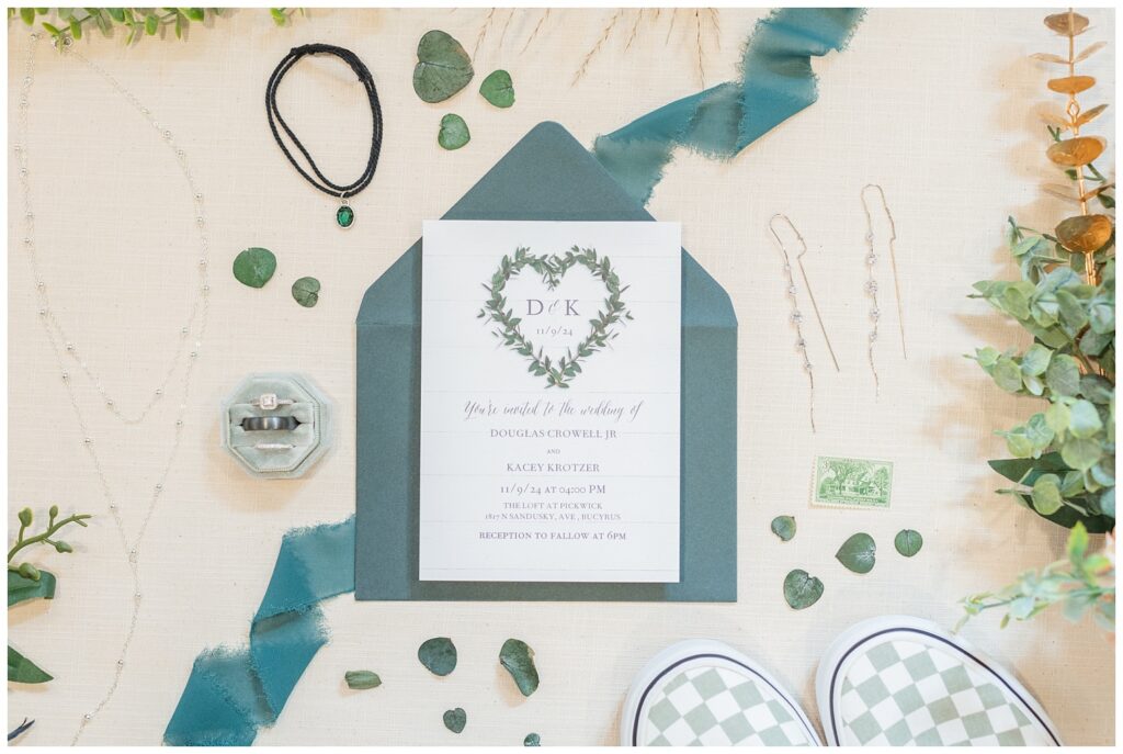 dark sage green invitation suite posed on a beige styling mat with wedding jewelry and flowers  