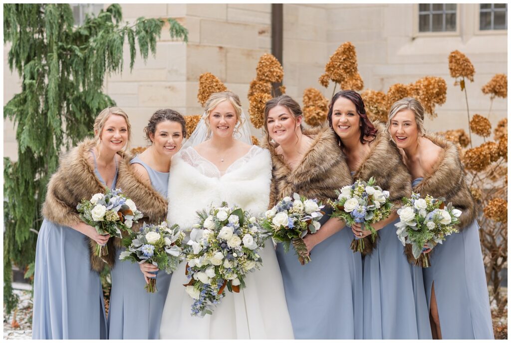 bride and bridesmaids wearing white and brown shawls and blue dresses outside 