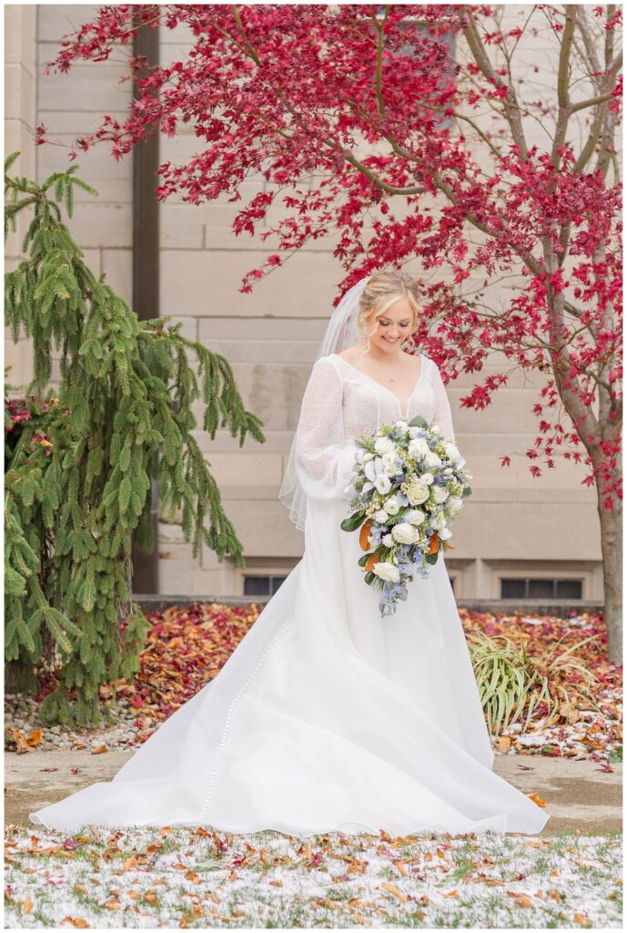 bride looking down at her bouquet in front of a large red tree before the ceremony