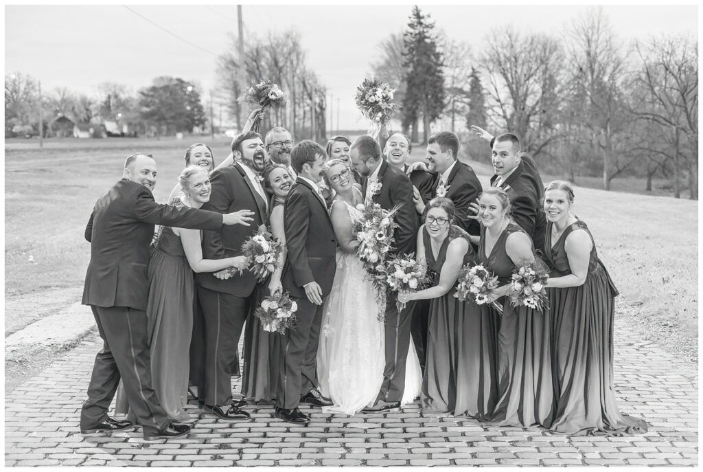 full wedding party crowding around the bride and groom on a brick road