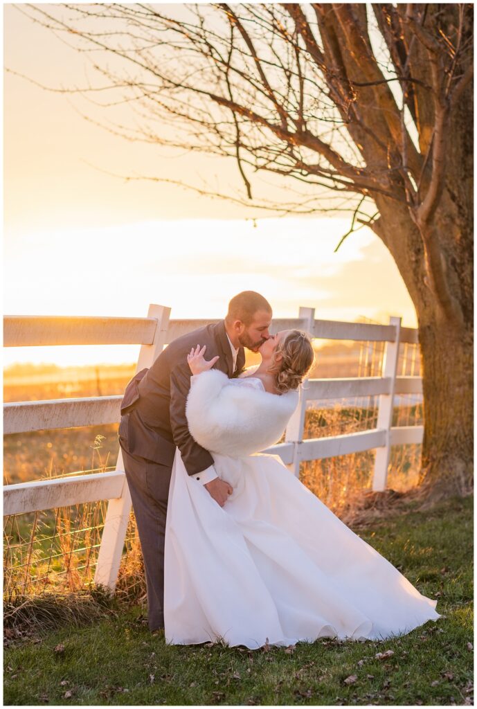 groom dipping the bride back for a kiss next to a white fence at sunset