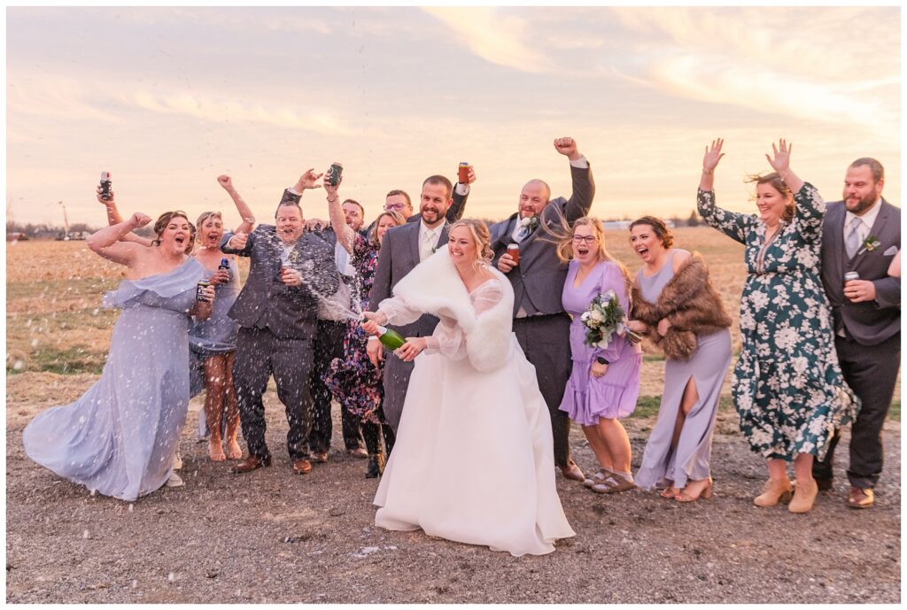 bride popping a bottle of champagne with family behind the groom's family barn