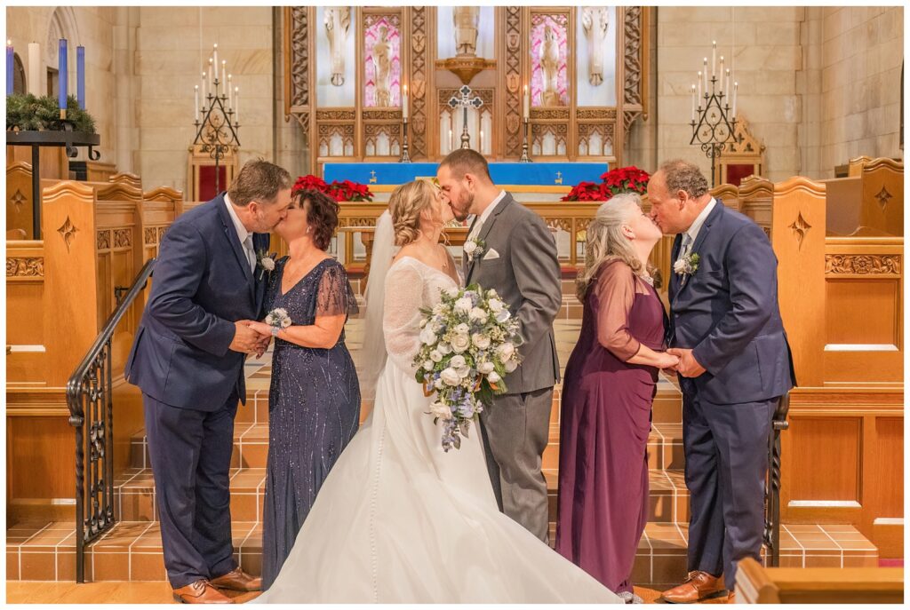 wedding couple, groom's parents and bride's parents all share a kiss at the church altar