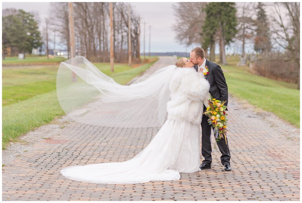 bride wearing a fur coat and kissing the groom with her veil waving in the wind 