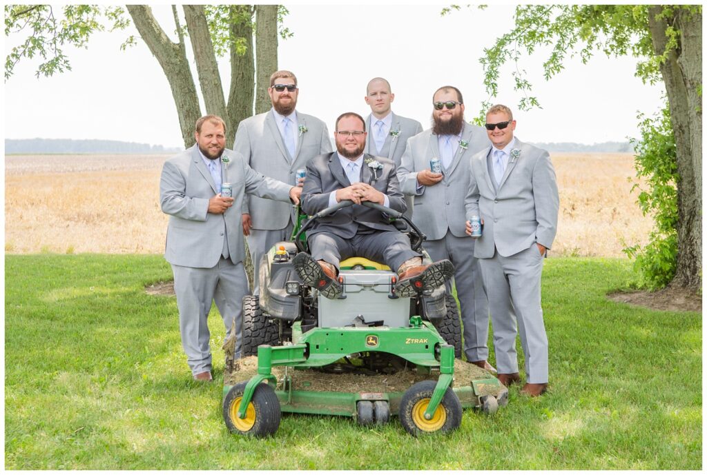 groom sitting on his green lawn mower with his groomsmen around him