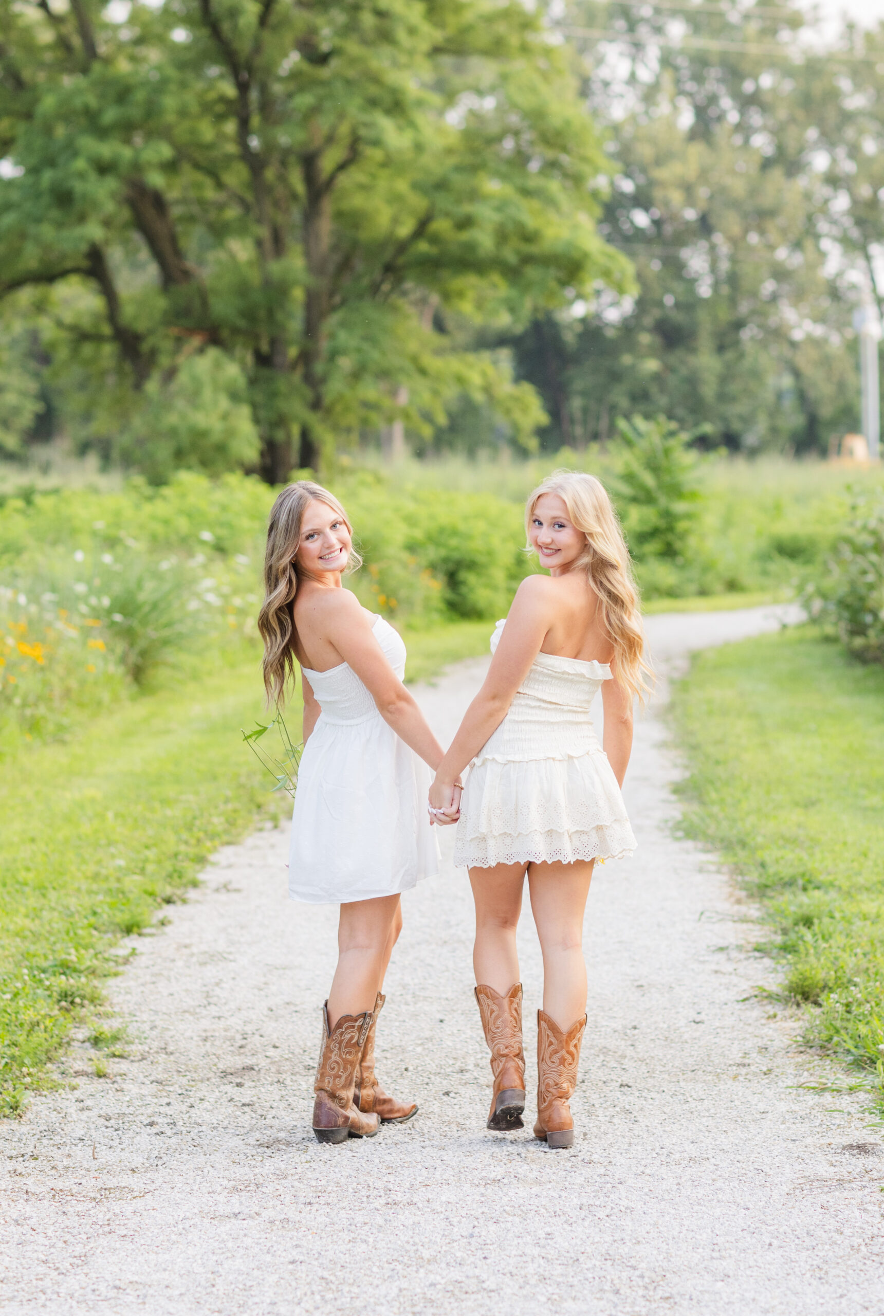 two seniors wearing white dresses and walking along a gravel road