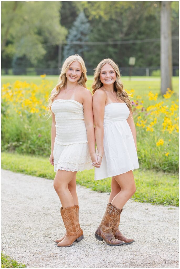 two senior best friends wearing white dresses and posing next to wildflowers